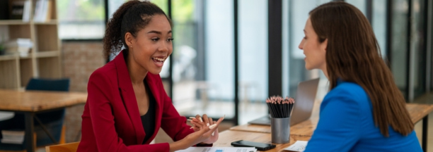 Two female employees discuss work facing each other at a table.