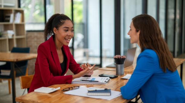 Two female employees discuss work facing each other at a table.