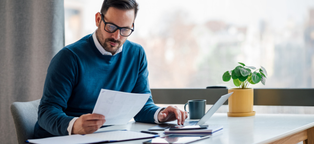 Man in blue sweater with glasses looks down at paper in his hand while sitting at a desk working.