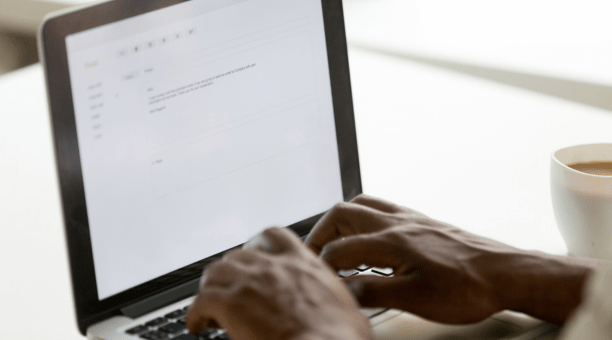 Hands of a person on a laptop keyboard writing an email with a cup of coffee next to them.