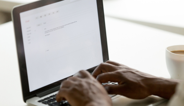 Hands of a person on a laptop keyboard writing an email with a cup of coffee next to them.