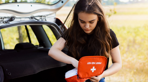 Young woman holding a first aid kit standing next to her car truck in front of a field.