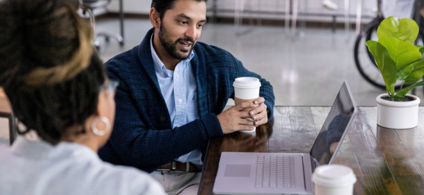 Two employees talk in front of open laptop while drinking coffee.