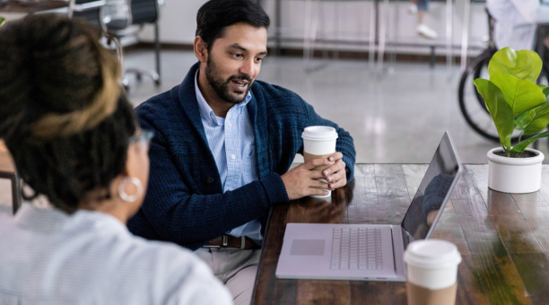 Two employees talk in front of open laptop while drinking coffee.