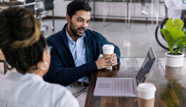 Two employees talk in front of open laptop while drinking coffee.