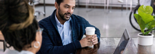 Two employees talk in front of open laptop while drinking coffee.